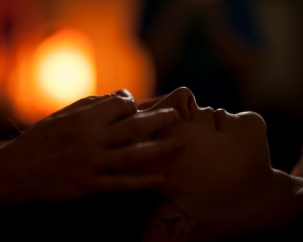 A practitioner's hands resting gently on a relaxed client's face in warm, low light